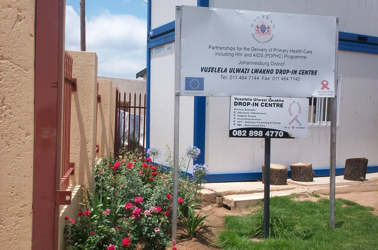 Vuselela drop-in centre signs beside a flower bed and a blue-and-white building.