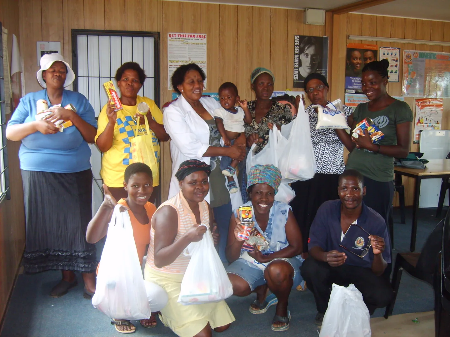 A group posing indoors, several holding grocery bags and packaged food, with a child near the centre.