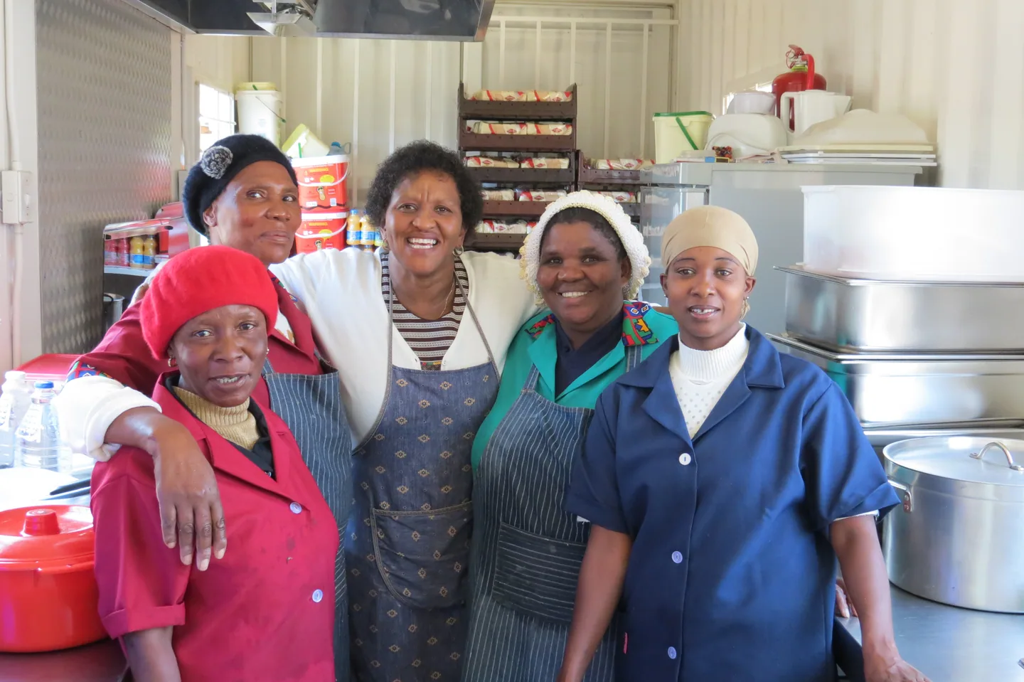 Five people standing together in a kitchen beside large cooking pots and food containers.
