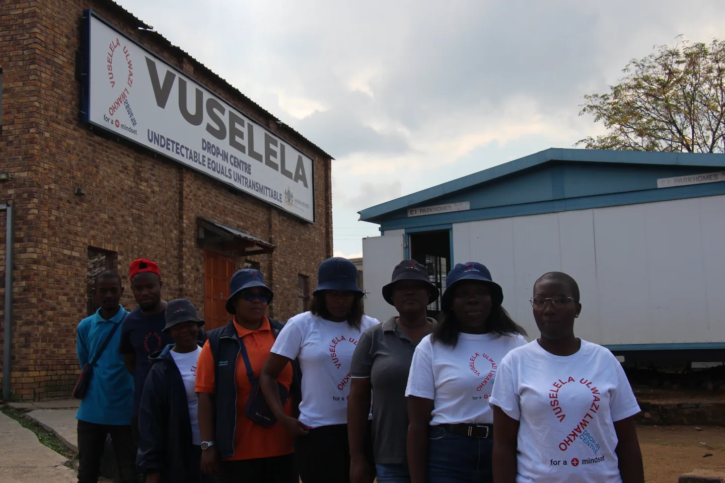 Eight people posing outside Vuselela, several wearing branded shirts and hats.